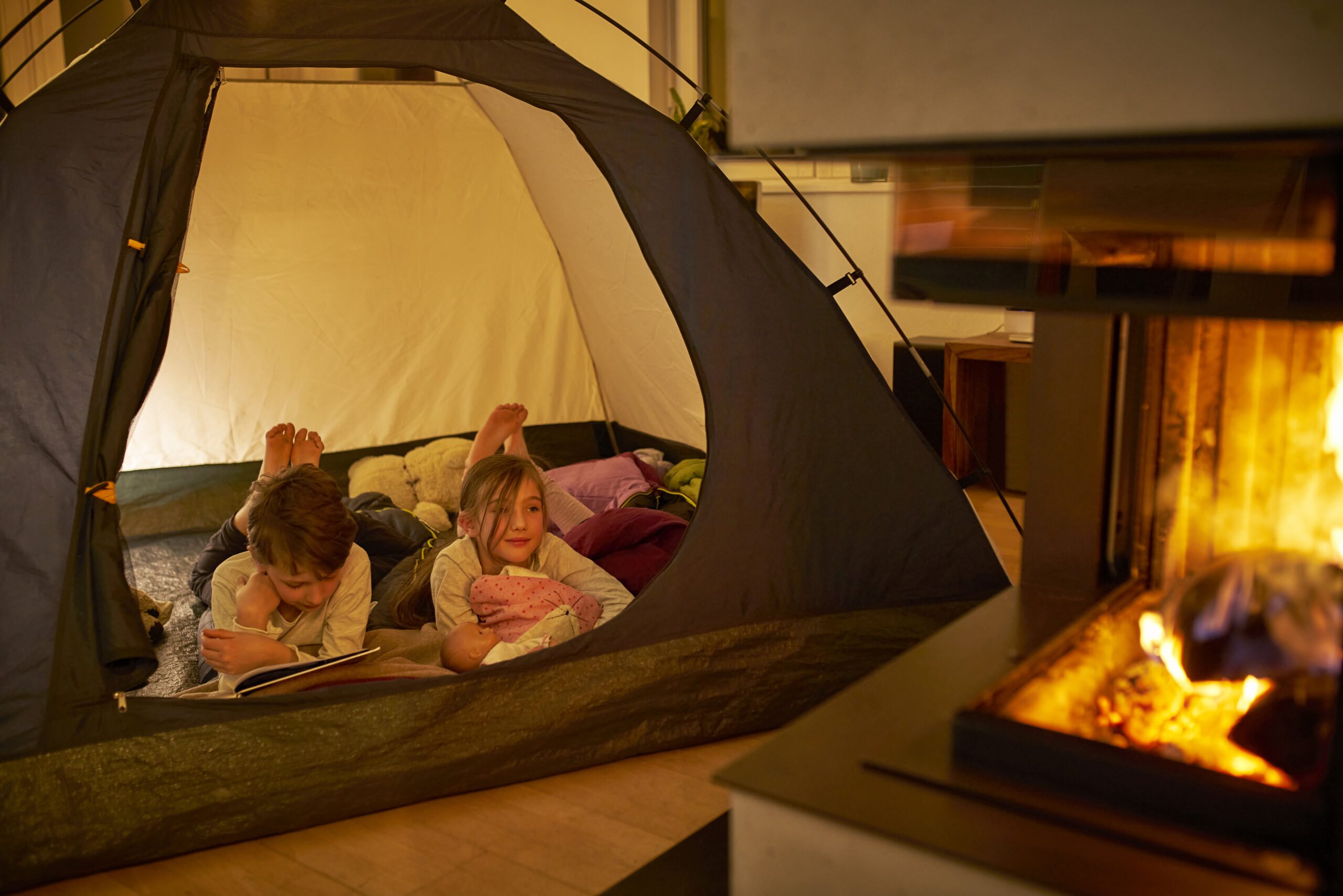 Kinder campen im Wohnzimmer wegen der Corona-Pandemie. Junge (10) liest seiner kleinen Schwester (8) vor. Feuer, Zelt.Zuhause bleiben.