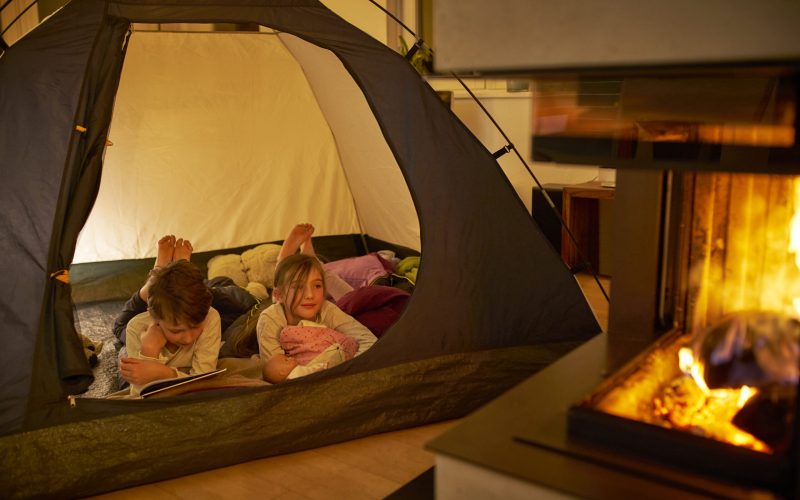 Kinder campen im Wohnzimmer wegen der Corona-Pandemie. Junge (10) liest seiner kleinen Schwester (8) vor. Feuer, Zelt.Zuhause bleiben.